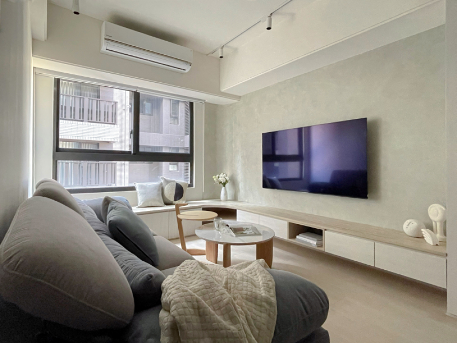 客廳系統櫃設計圓角造型,將電視櫃延伸與窗邊臥榻做結合,可作為親友來訪時的座席,同時也可增加收納量。電視牆使用低飽和度的灰色塗料,使整體空間帶有天然材質且明亮