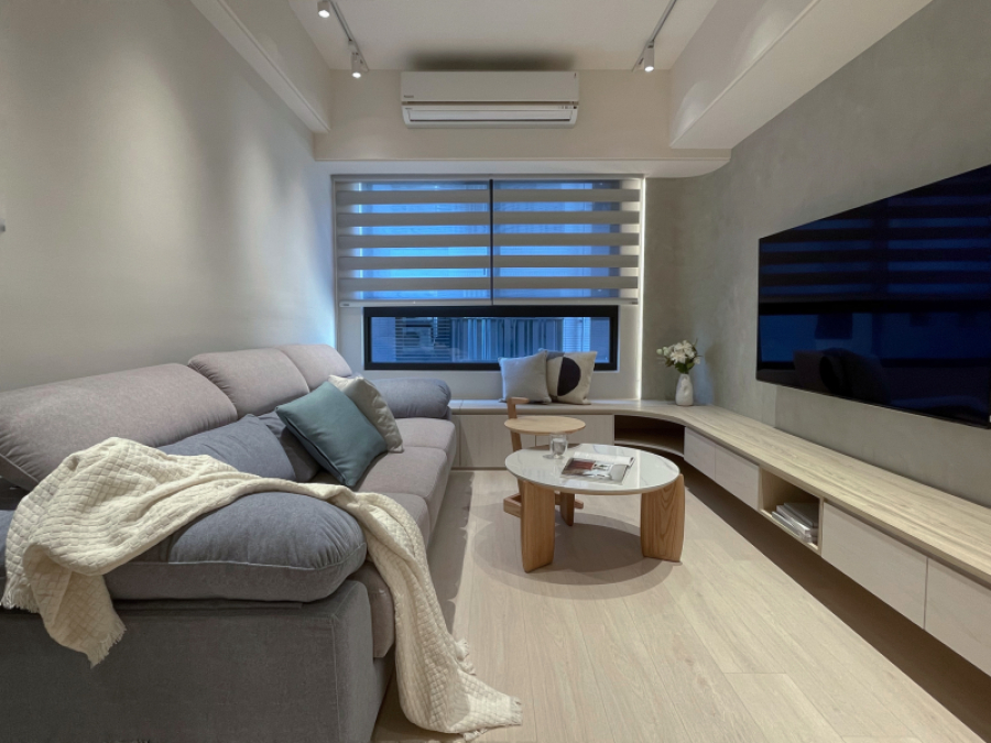 客廳系統櫃設計圓角造型,將電視櫃延伸與窗邊臥榻做結合,可作為親友來訪時的座席,同時也可增加收納量。電視牆使用低飽和度的灰色塗料,使整體空間帶有天然材質且明亮